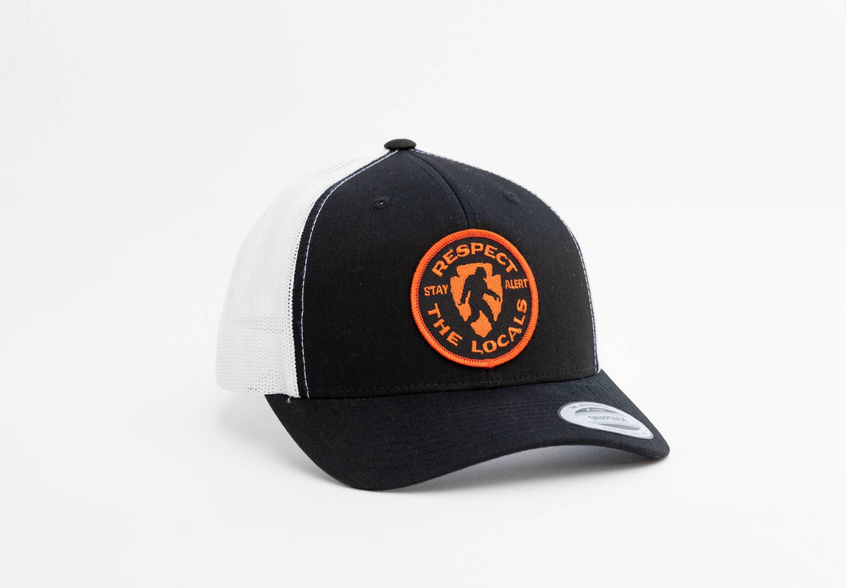 Black crown and white mesh meet on the Bigfoot Respect The Locals trucker hat with its orange circular patch.