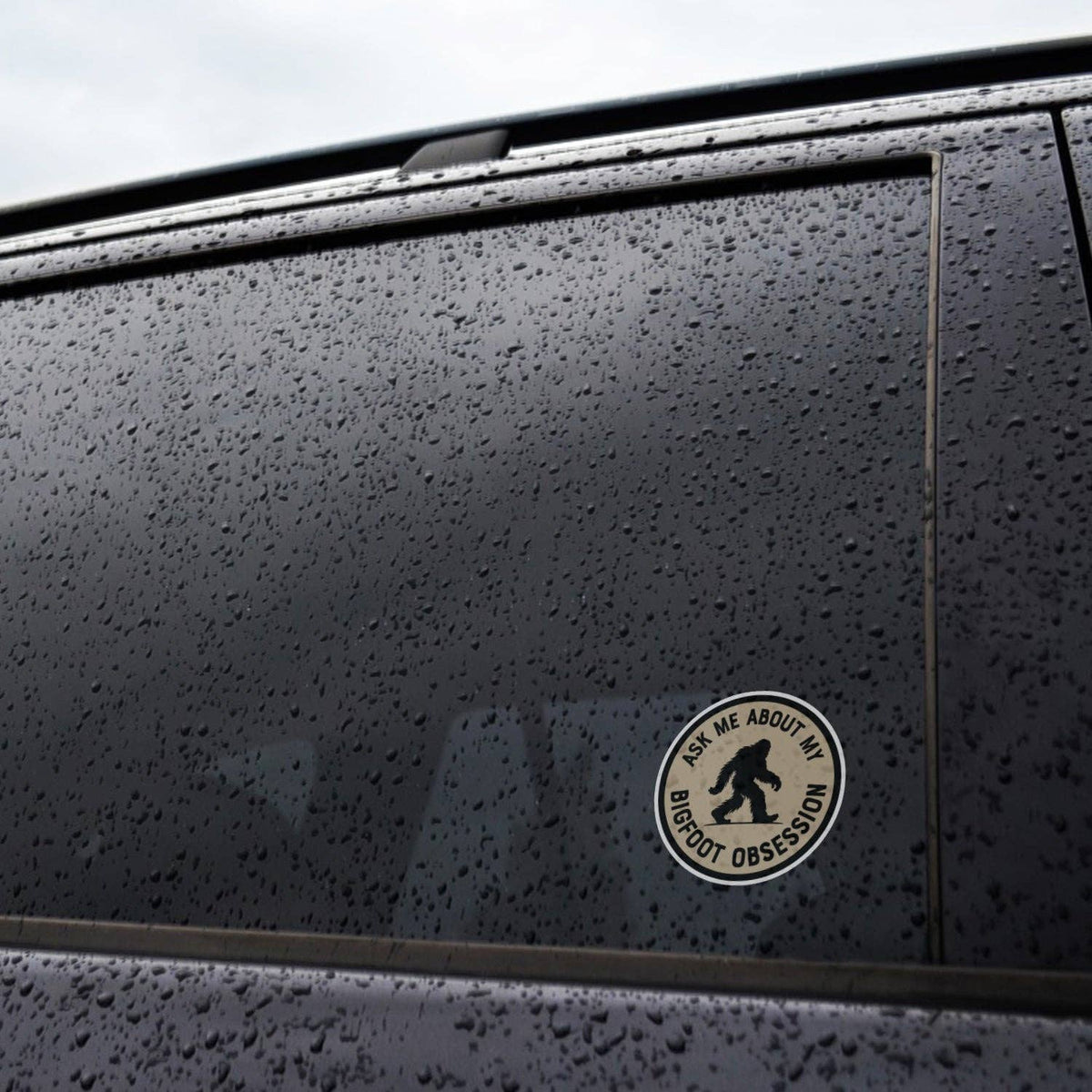 Stuck to the rain beaded window of a dark car, the Ask Me About My Bigfoot Obsession sticker catches the light.