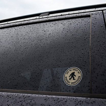 Stuck to the rain beaded window of a dark car, the Ask Me About My Bigfoot Obsession sticker catches the light.