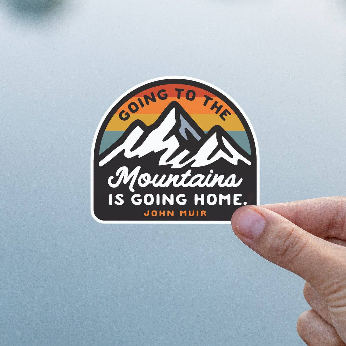 Against a soft grey blue backdrop, a thumb and finger grip the Going to the Mountains is Going Home sticker.