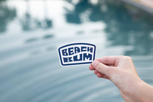 A hand holds the navy Beach Bum sticker above rippling pool water lit by afternoon sun.