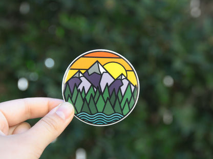 Small white bokeh dots float behind the Mountains Call sticker as fingers hold it away from the hedge.