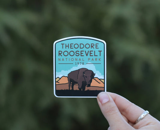 A hand holds the Theodore Roosevelt National Park sticker with its grazing bison against blurred green branches.