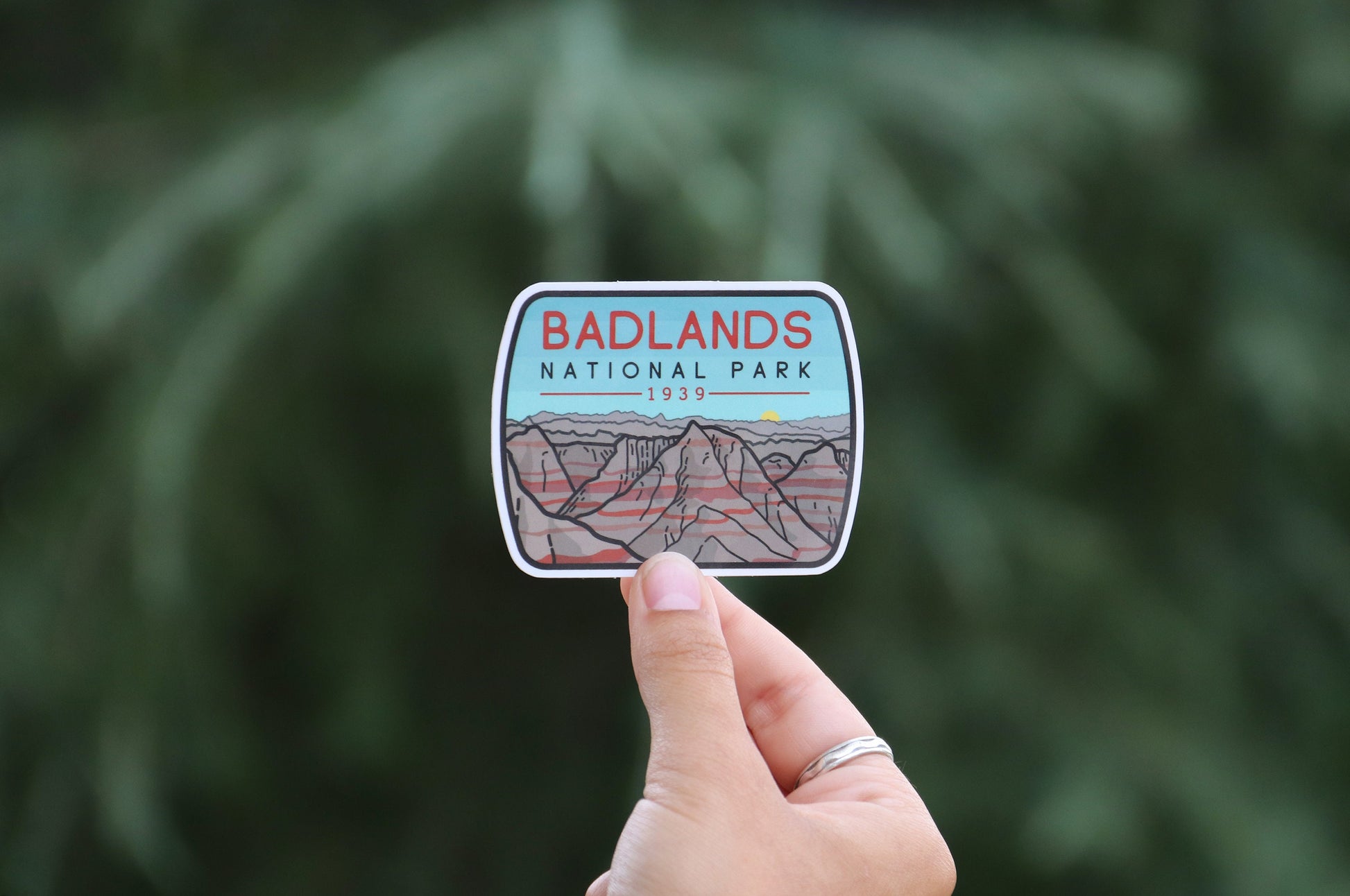 Pulled back a little, the Badlands National Park sticker sits small in a ringed hand before a pine tree.