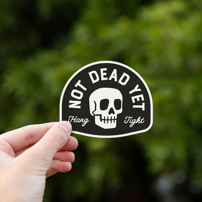 Someone holds the Not Dead Yet sticker up against a blurred backdrop of dark green leaves.