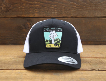Straight on against dark barn wood, the black and white Yellowstone National Park trucker hat sits on planks.