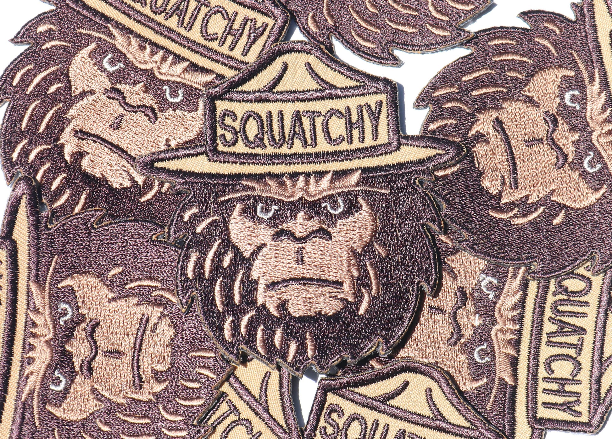 A close pile of Squatchy patches overlaps, their embroidered beards and lettering running in every direction.