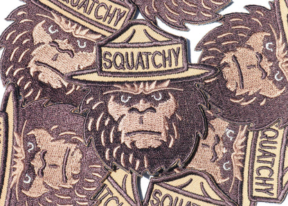 A close pile of Squatchy patches overlaps, their embroidered beards and lettering running in every direction.