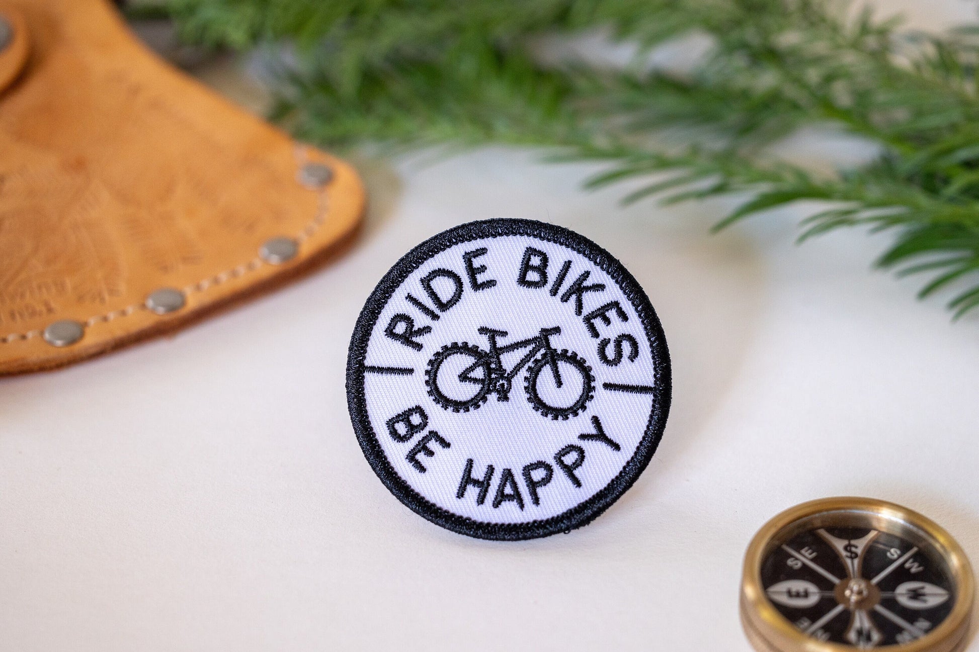 Beside a brass compass and a leather cuff, the Ride Bikes Be Happy patch rests near a sprig of cedar.
