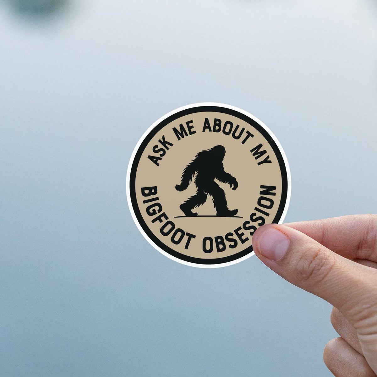 Thumb and finger grip the Ask Me About My Bigfoot Obsession sticker against a hazy blue backdrop.