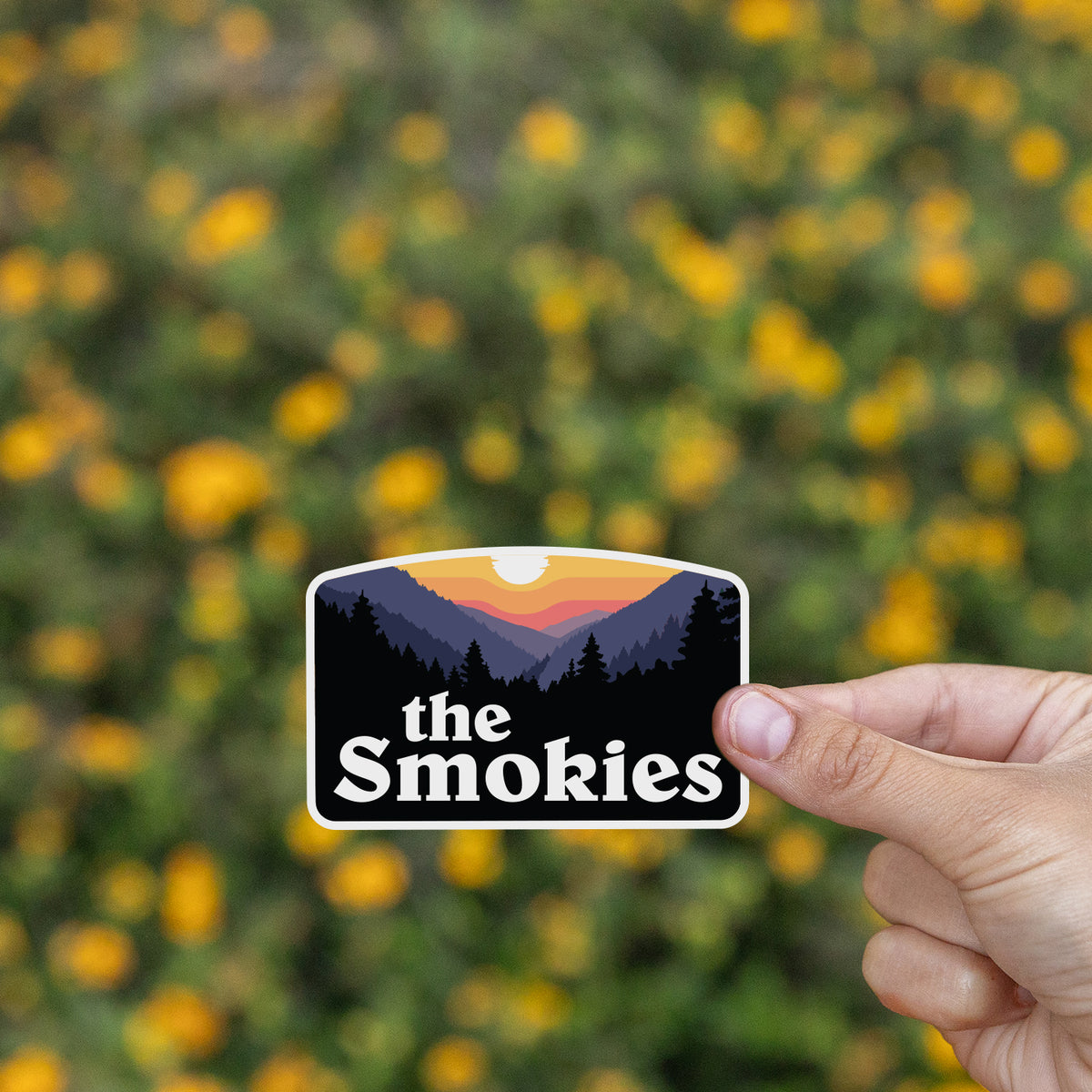 A hand presents the The Smokies sticker in front of a blurred field of small yellow wildflowers.