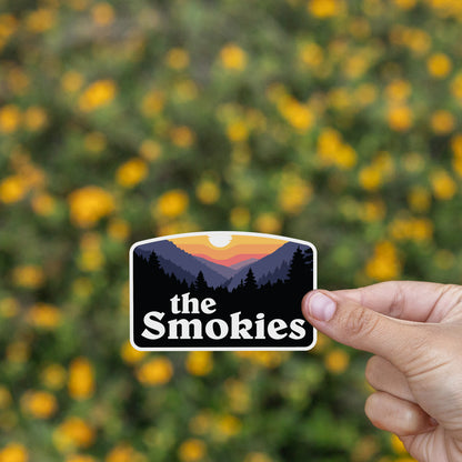A hand presents the The Smokies sticker in front of a blurred field of small yellow wildflowers.