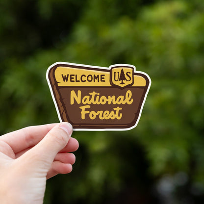 Fingers grip the National Forest Sign sticker against a blur of bright green leaves.