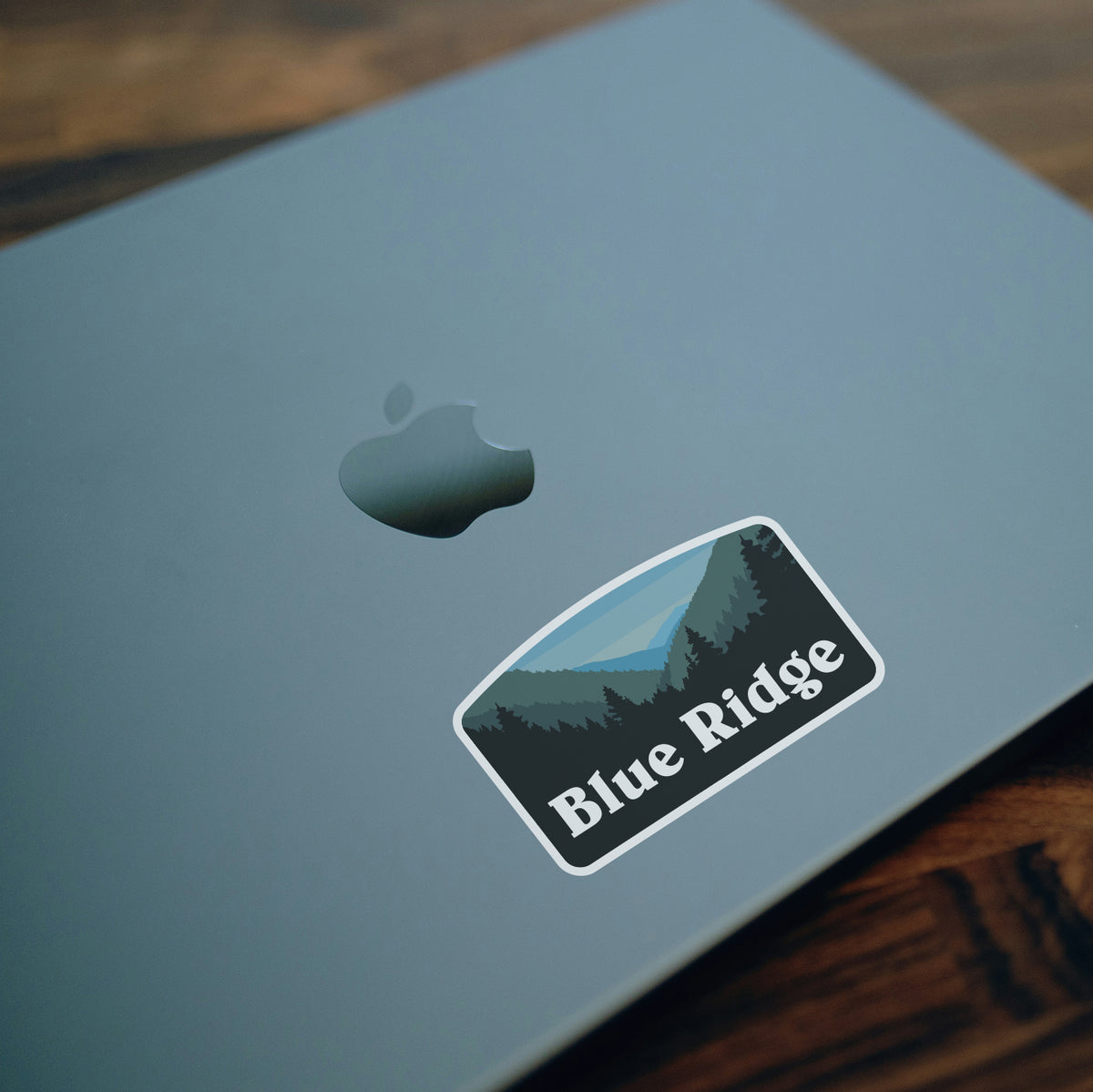 Stuck below the Apple logo on a gray laptop lid, the Blue Ridge sticker rests on a wooden desk.