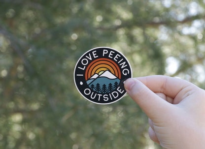 Sunlight through tree branches backs the I Love Peeing Outside sticker held in a raised hand.