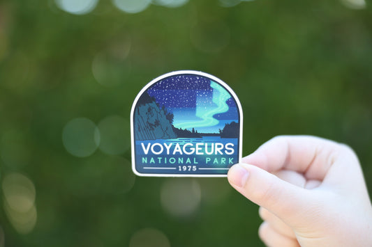 Pulled back against blurred green leaves, the Voyageurs National Park sticker shows its full arched shape.