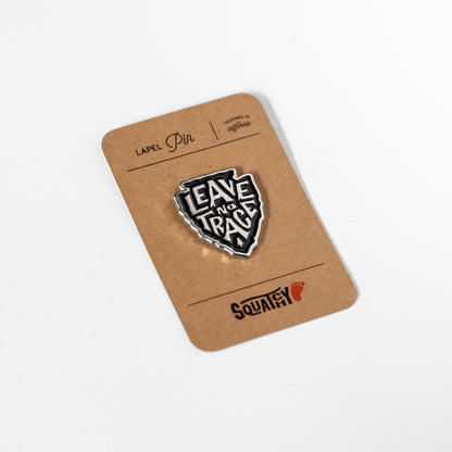 Resting at an angle on its kraft backing card, the Leave No Trace pin shows the Squatchy footprint stamp below.