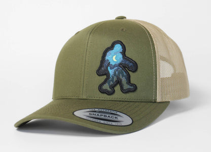 Olive and khaki Bigfoot Night Sky trucker hat facing front, its starry blue Sasquatch patch centered above the brim.
