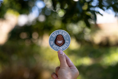 Fingers hold the Believe in Yourself sticker up against blurred green leaves and afternoon sun.