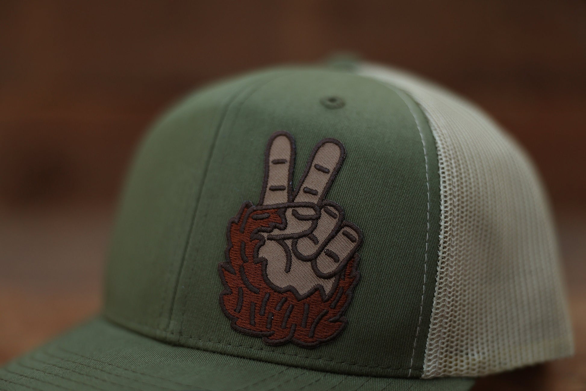 Close on the green crown, the Bigfoot Peace Left Side Trucker Hat patch shows two fingers rising from shaggy brown fur.