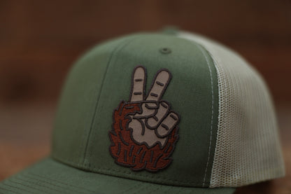 Close on the green crown, the Bigfoot Peace Left Side Trucker Hat patch shows two fingers rising from shaggy brown fur.