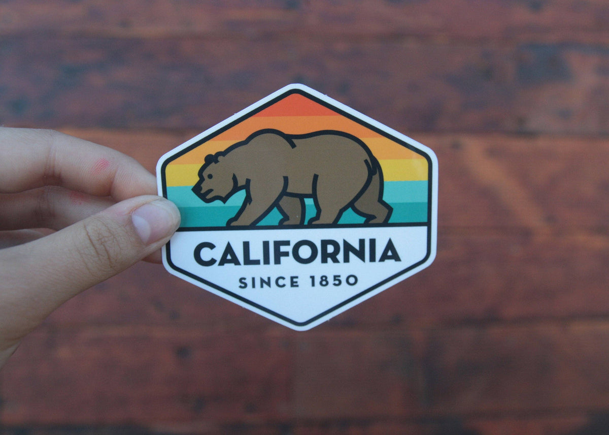 Warm red brick sits behind fingers gripping the edge of the California State Bear sticker.