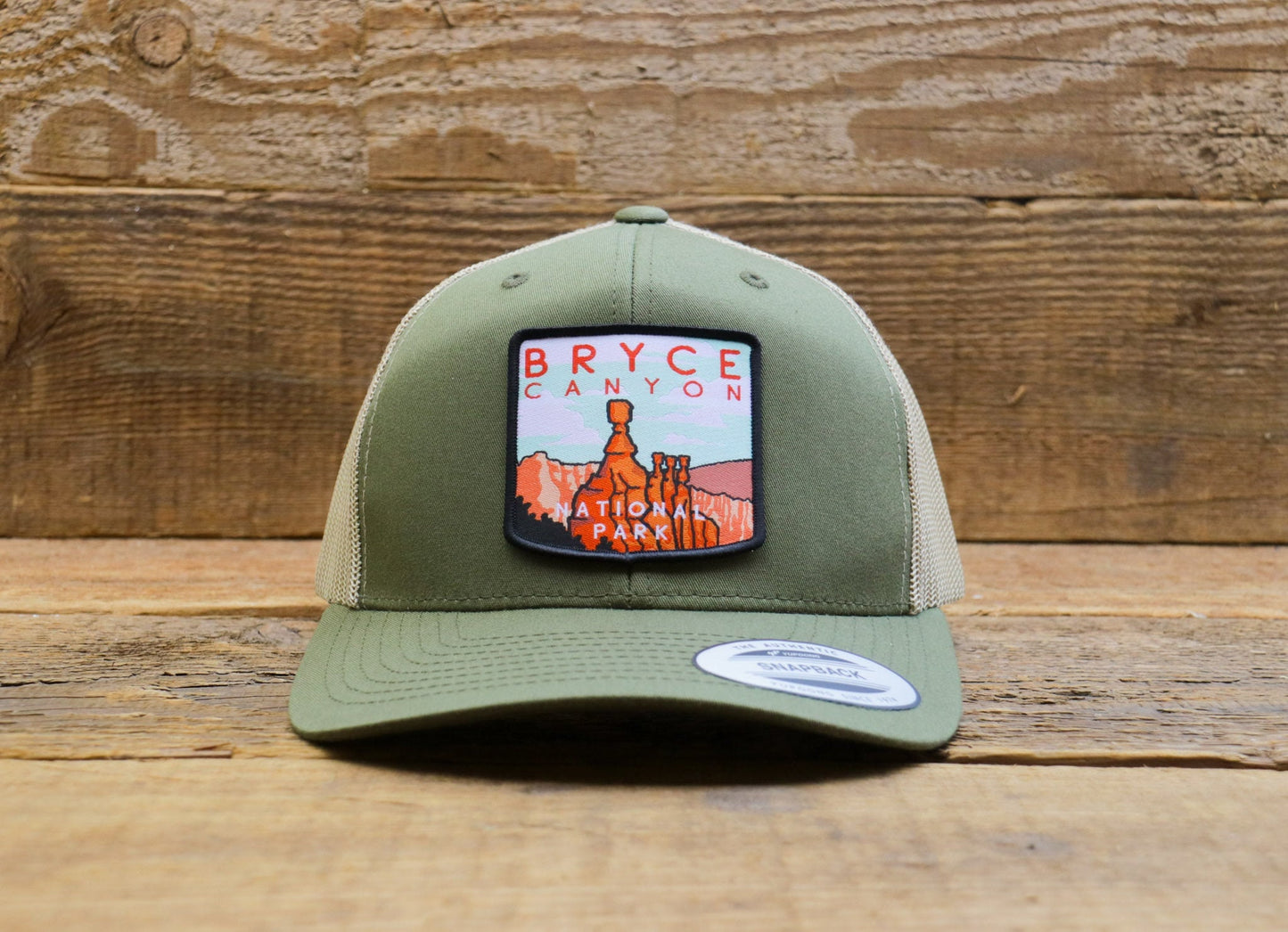 Straight on before barn boards, the Bryce Canyon National Park trucker hat centers its hoodoo and red lettering patch.