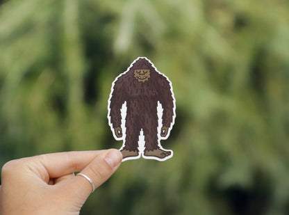 Held a little lower and closer, the Happy Bigfoot sticker shows its smiling face before soft pine branches.