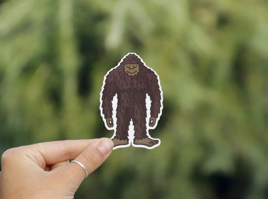 Held a little lower and closer, the Happy Bigfoot sticker shows its smiling face before soft pine branches.