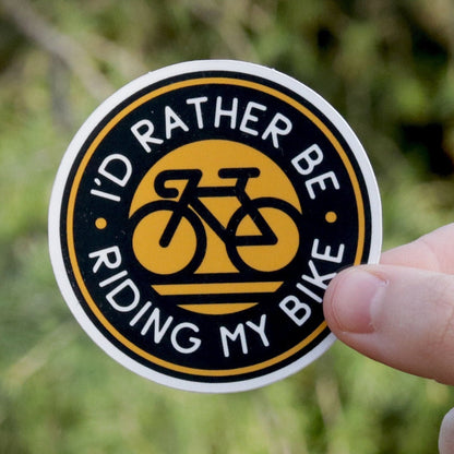 Held further back at arm's length, the I'd Rather Be Riding My Bike sticker sits small against sunlit branches.