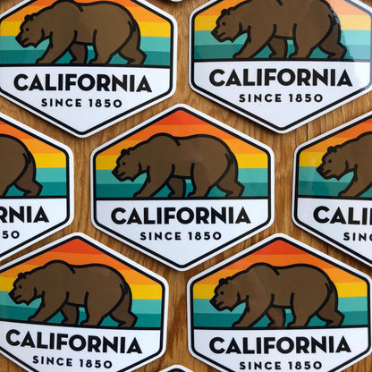 Overlapping rows of California State Bear stickers cover a wooden tabletop.