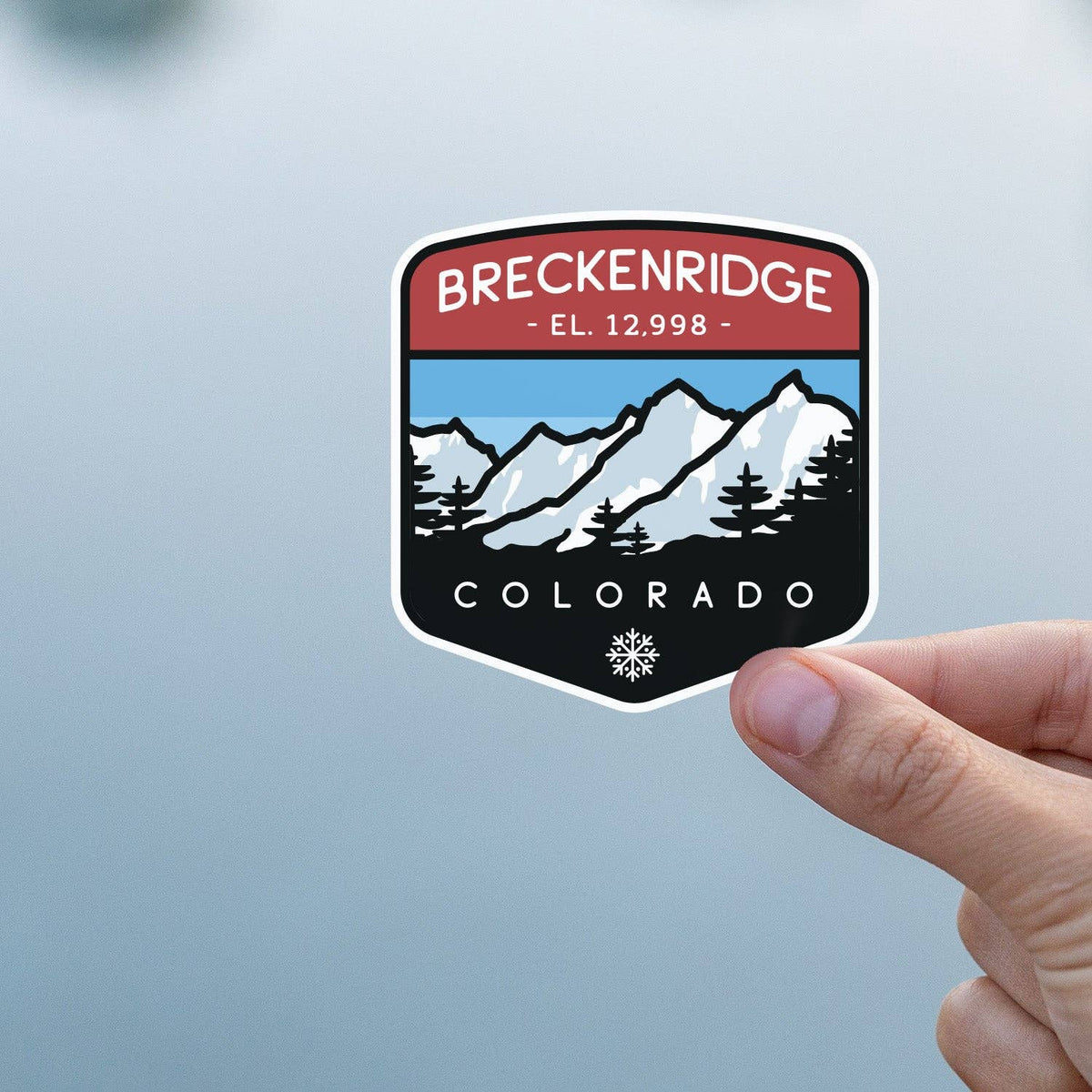 Against a pale grey wall, a thumb and finger steady the Breckenridge Colorado sticker.