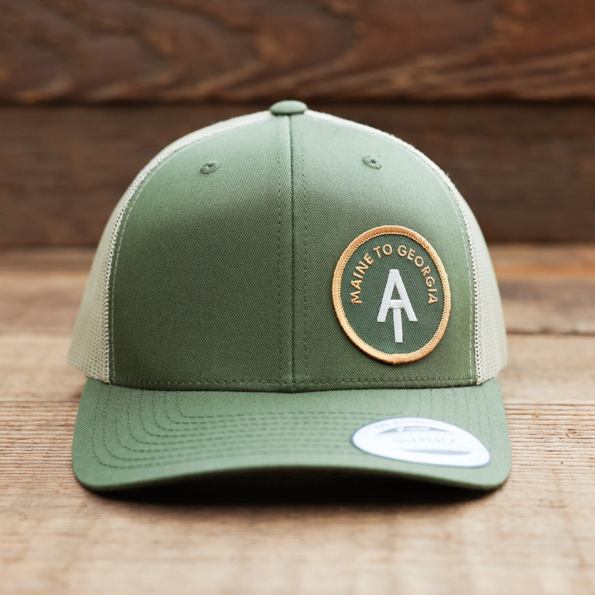 Standing on weathered planks, the green Appalachian Trail trucker hat faces forward with a sticker on the brim.