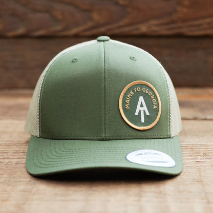 Standing on weathered planks, the green Appalachian Trail trucker hat faces forward with a sticker on the brim.