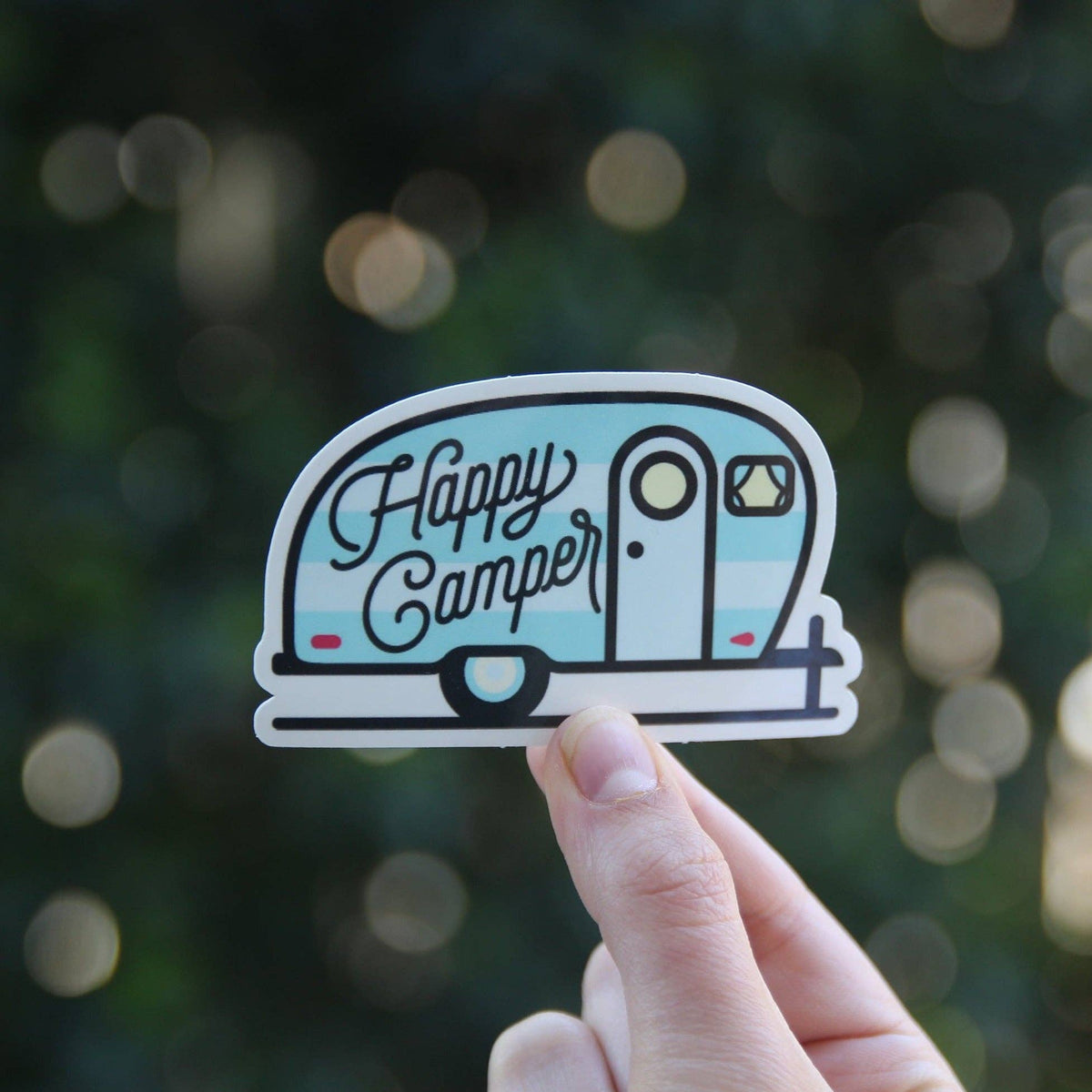 Fingers hold up the Happy Camper sticker, its blue trailer and script glowing against soft green bokeh.