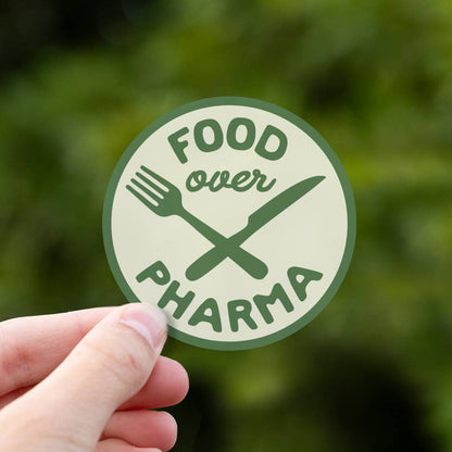 A hand holds the round Food over Pharma sticker, its crossed fork and knife green against blurred leaves.