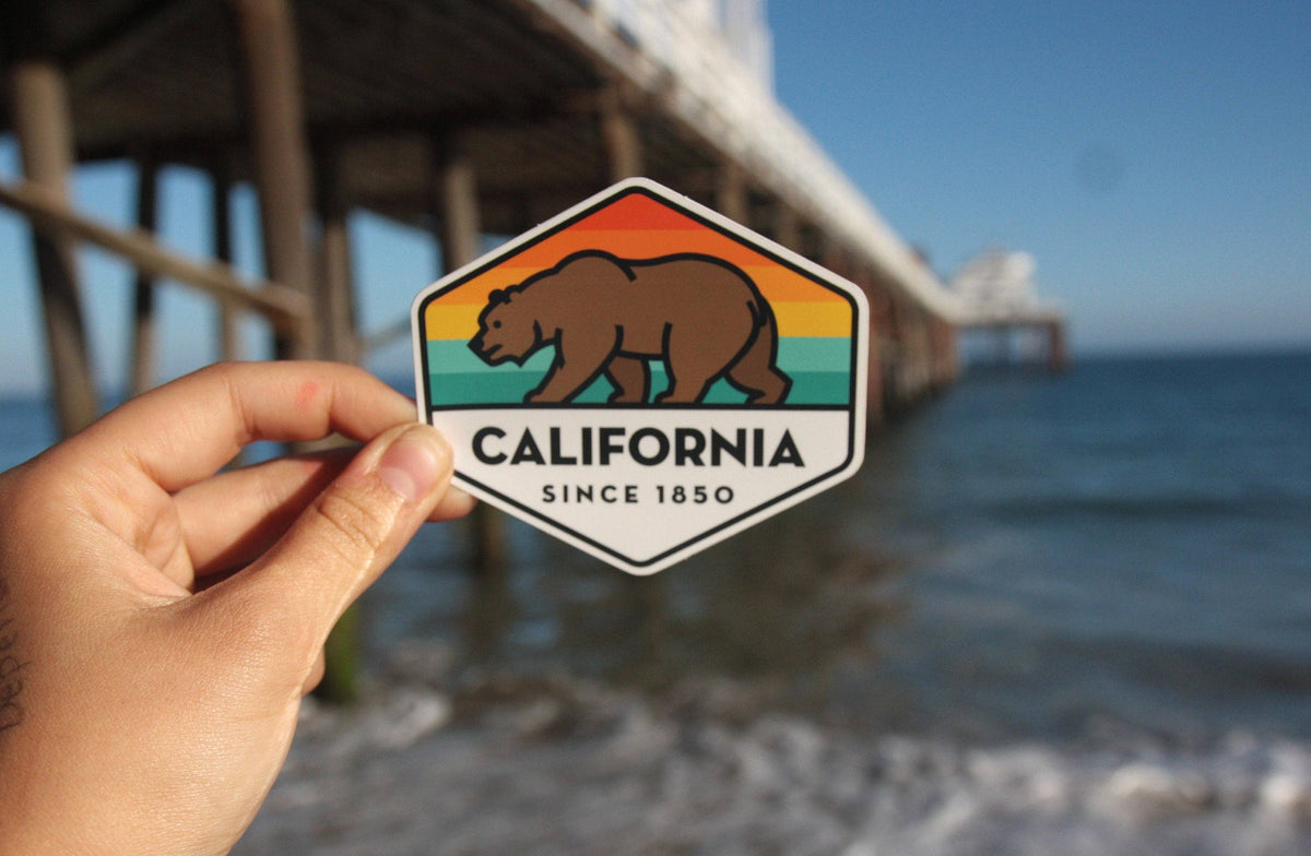 Beneath a long wooden pier, the California State Bear sticker is held above the surf.