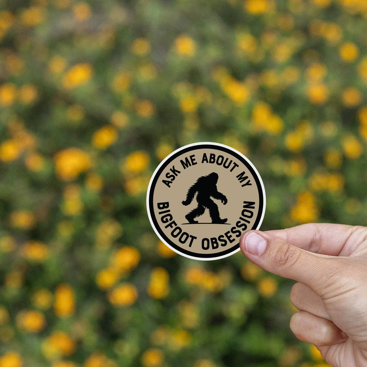 Yellow wildflowers blur behind fingers holding the Ask Me About My Bigfoot Obsession sticker.