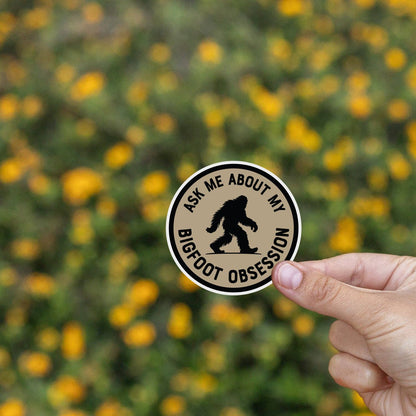 Yellow wildflowers blur behind fingers holding the Ask Me About My Bigfoot Obsession sticker.