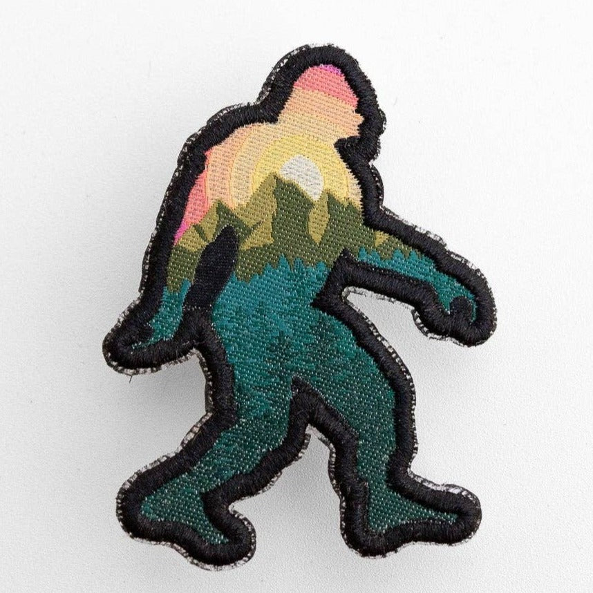 On a plain white surface, the Bigfoot Adventure Patch shows a sunset and pine forest inside its black outline.