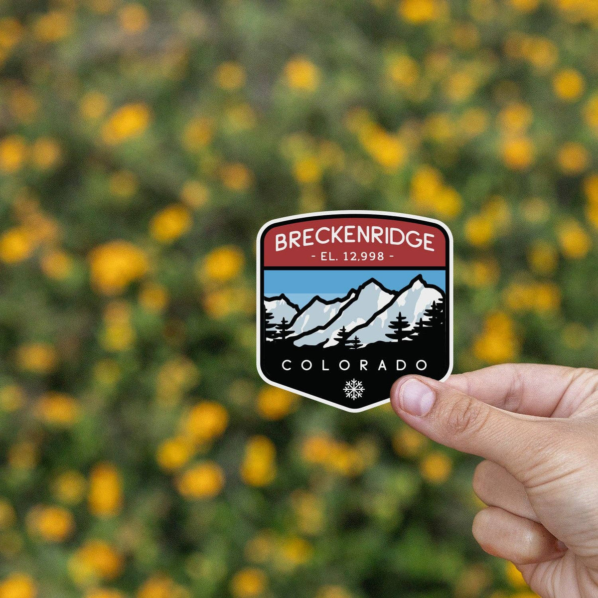 Yellow wildflowers blur behind a hand pinching the Breckenridge Colorado sticker.