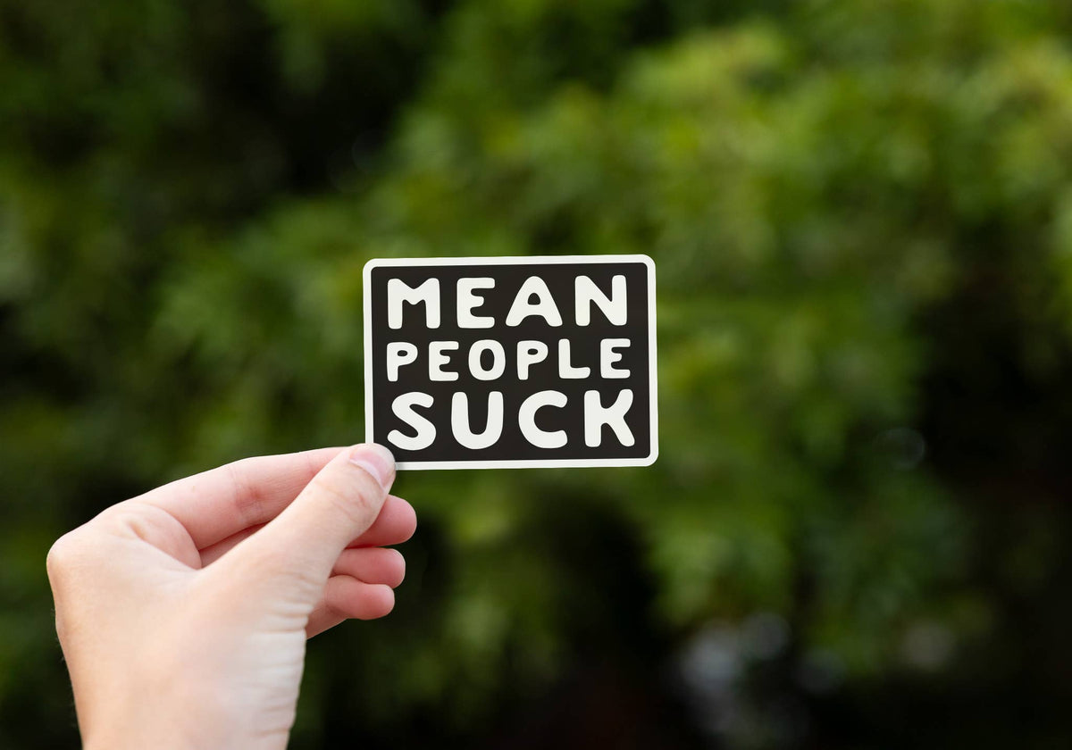 A hand holds the Mean People Suck sticker up against blurred green foliage in daylight.