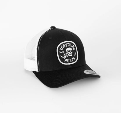 Black and white Everything Hurts trucker hat sits angled, skull patch centered above the curved black brim.