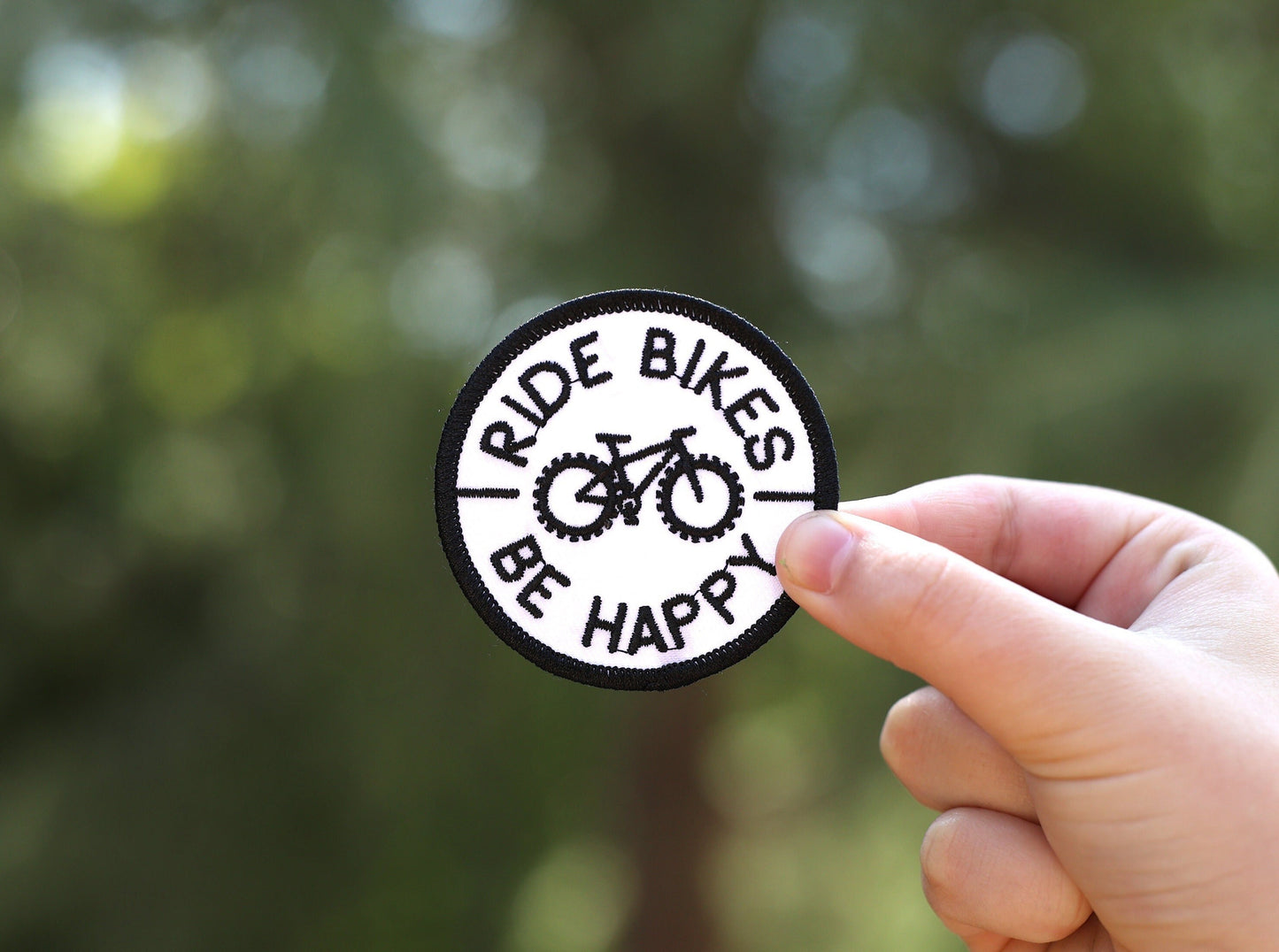 Held between finger and thumb in daylight, the Ride Bikes Be Happy patch stands against soft green bokeh.