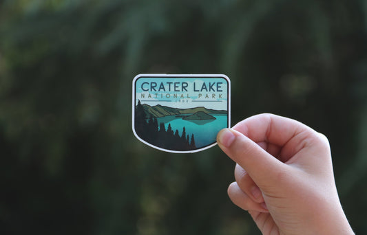 Held farther back against green conifer branches, the Crater Lake National Park sticker shows its full arched shape.