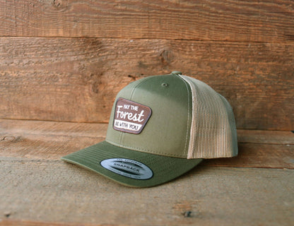 Standing on weathered boards, the green May The Forest Be With You trucker hat rests against a barn wood wall.