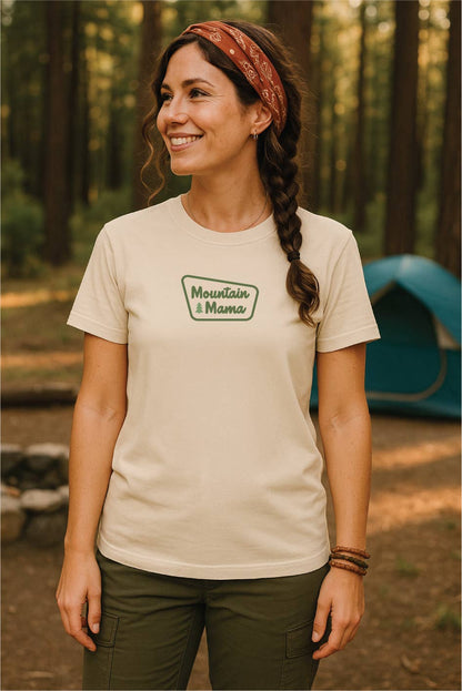 Standing in a pine campground near a teal tent, a woman wears the ivory Mountain Mama T shirt with a braid.
