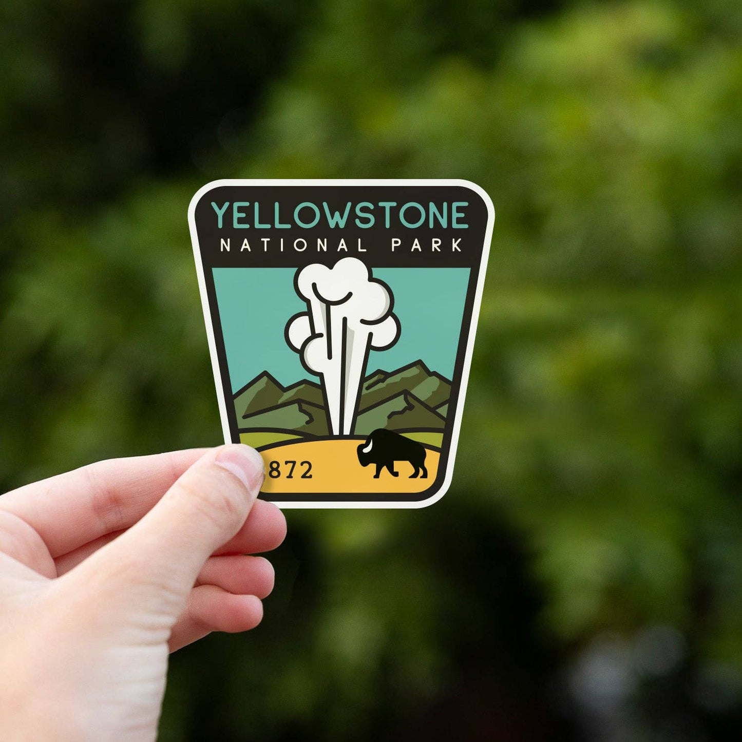 A hand holds the Yellowstone National Park sticker up against blurred green foliage in daylight.