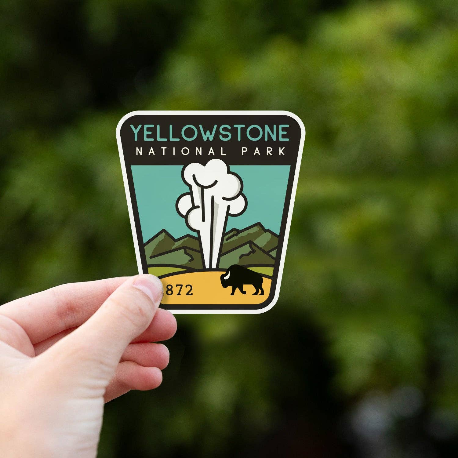 A hand holds the Yellowstone National Park sticker up against blurred green foliage in daylight.
