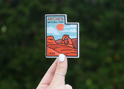 Held further from the camera, the Arches National Park sticker sits small against a wide hedge of green.